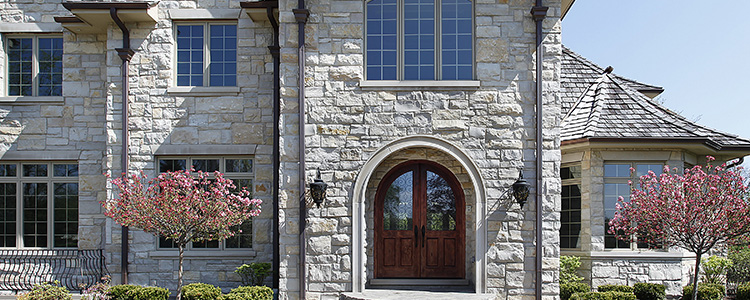Replacement windows in a large stone house with arched wooden double front doors and pink flowering trees in front.