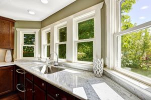 Kitchen cabinet with marble top and sink with row of white double-hung windows over counter.